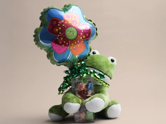 A plush green frog toy with white paws sits on a neutral beige surface, holding a small clear plastic container filled with assorted colorful candies. A shiny, multicolored foil balloon shaped like a flower with blue, pink, and orange petals is attached to the frog with a green curly ribbon. The composition is playful and gift-like, set against a simple background.